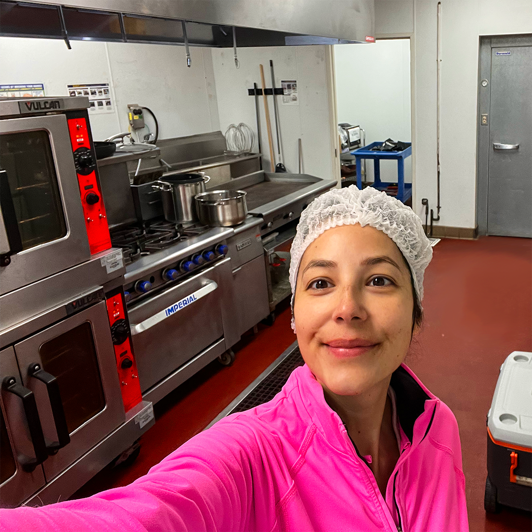 Person in a kitchen wearing a pink jacket and hairnet, standing in front of industrial cooking equipment.